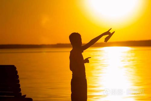 小男孩在日落时站在河的背景上他手里拿着一架纸飞机用手指指着太阳