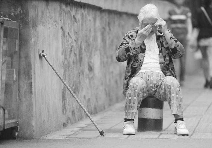 一位独自散步的老人走累了,把拐杖靠在墙边,坐在隔离桩上,擦了擦