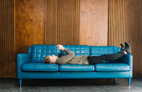 一个年轻人躺在一个时髦的蓝色沙发上, 使用智能手机.