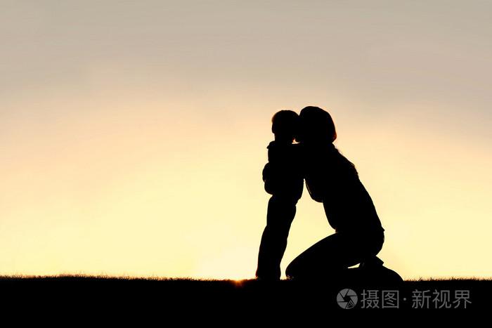 年轻母亲拥抱蹒跚学步的儿子在日落时的剪影