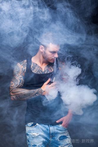 那个带着纹身的肌肉发达的男人在黑暗的背景上抽着一支电子香烟.
