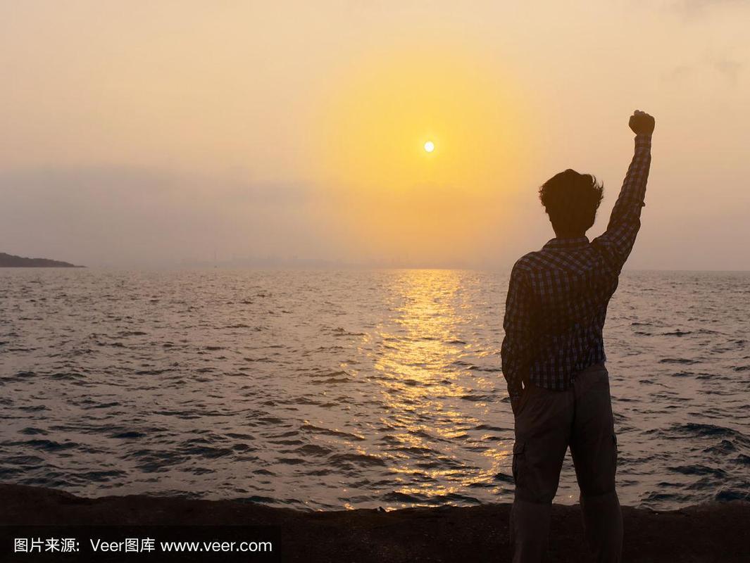 放松的年轻亚洲男子在海边举起手与日落背景的背影.